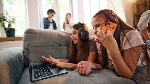 Girls multitasking between smartphone conversation and laptop at home with friends in the background. Concept of cross-device behavior, connected social life, and digital communication.
