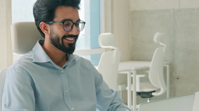 Cheerful happy Indian Arabian business man millennial entrepreneur working online on laptop inside office typing email chatting client smile male manager businessman programmer guy message computer