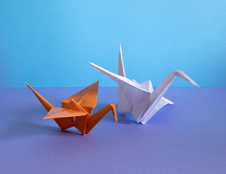 Two origami cranes, one orange and one white, rest on a purple surface with a blue background, symbolizing peace, balance, and creative expression.