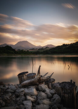 View of Lago di Cingoli at sunset with tree stumps and rocks in the foreground, mountains in the distance, and soft clouds in the sky, Cingoli, Marche, Italy.