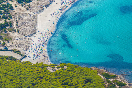 Aerial view of a crowded beach with turquoise water, colorful umbrellas, and lush pine forests along the coast in Sardinia, Italy.