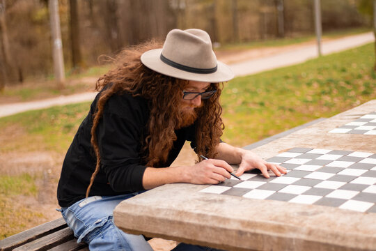 Bearded Hippie Man Wearing Hat Drawing On Checkerboard Table Outdoors