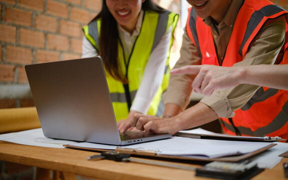 Team of Asian engineers collaborating on blueprints and a laptop inside a construction site.