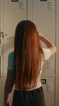 Blonde Schoolgirl Organizes Decorated Locker, Reaching in to Grab Pink Notebook before Class, while Brunette Friend Stands Beside in School Corridor. Education, Daily School Routine. Vertical Shot.