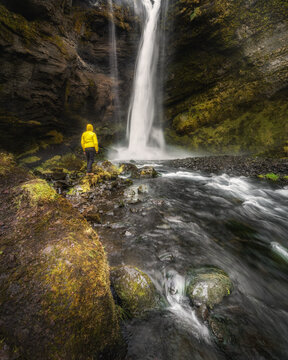 View of a person in a yellow jacket standing before the cascading Kvernufoss waterfall, surrounded by mossy rock walls and a flowing river in Evindarholar, Rangarthing eystra, Iceland.