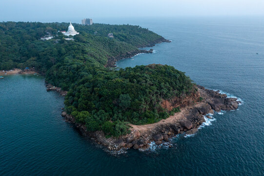 Aerial perspective of a densely forested rocky headland jutting out into the calm blue sea, with a majestic white peace pagoda visible in the background.