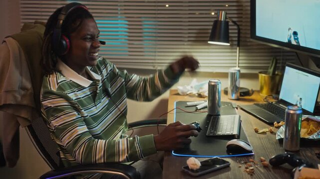 Young adult African American man playing video game with controller, celebrating victory with raised fists and leaning back in chair. Clip is suitable for gaming ads or tech addiction campaigns