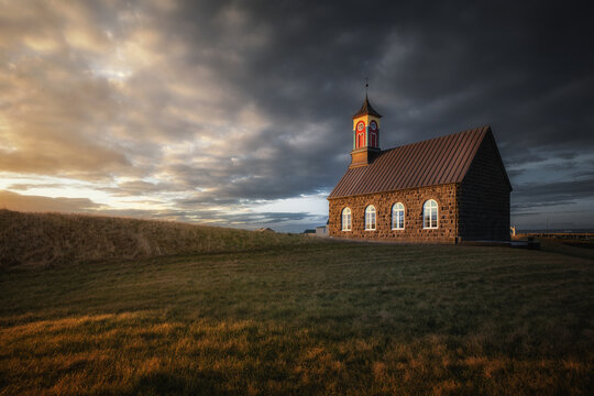 View of a historic stone church with a colorful steeple standing in a grassy field under a dramatic sunset sky in Grindavikurbaer, Iceland.