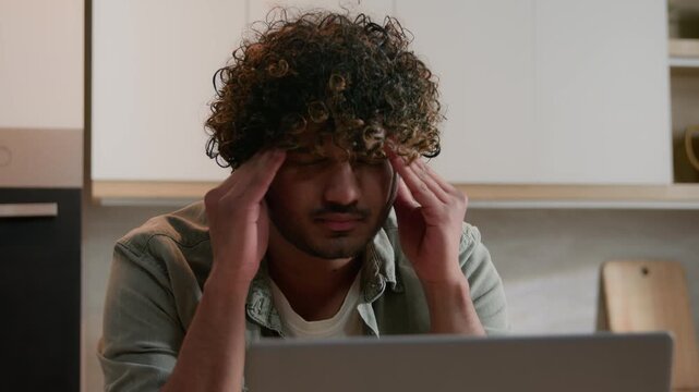 Tired Indian freelancer business man with headache using laptop at home kitchen exhausted Arabian male businessman reading bad news suffering migraine overwork stress failure computer problem indoor