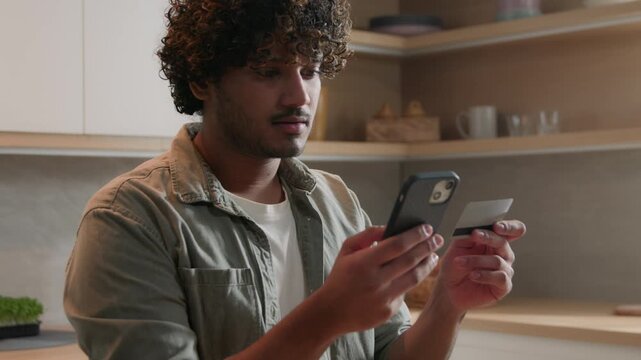 Indian Arabian man ordering food online with smartphone and credit card at home kitchen shopping grocery delivery app ethnic male guy customer mobile purchase secure payment ecommerce buying products