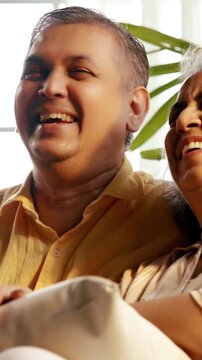 Indian Asian couple in their fifties with grey hair sitting on a sofa, enjoying an evening TV show and laughing together, illuminated by cozy evening light