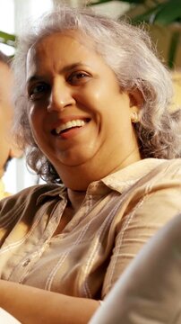 Indian Asian couple in their fifties with grey hair sitting on a sofa, enjoying an evening TV show and laughing together, illuminated by cozy evening light