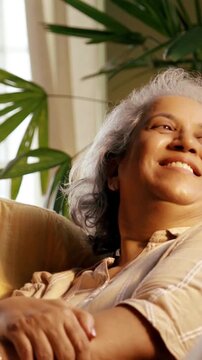Indian Asian couple in their fifties with grey hair sitting on a sofa, enjoying an evening TV show and laughing together, illuminated by cozy evening light
