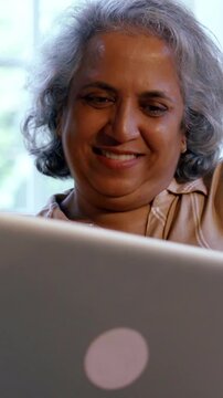 Indian Asian couple in their fifties with gray hair making a video call on laptop or smartphone, sitting on a sofa near a sunlit window at home, enjoying the moment