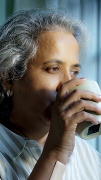 Indian Asian couple in their fifties with grey hair standing by a sunlit window, holding coffee or tea mugs, sharing a moment of quiet with pleasant expressions