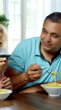 Indian Asian couple in their fifties, enjoying homemade noodles together in cozy dining interiors, sharing laughter and good times while savoring their meal from a bowl.