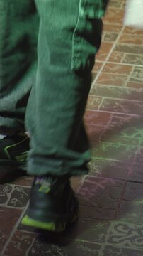 A person in cargo pants and sneakers dances rhythmically on a tiled floor illuminated by changing neon club lights.