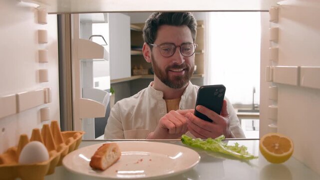 Hungry Caucasian man opening empty refrigerator idea food solution ordering online delivery mobile phone app POV inside fridge confused male guy seeing lemon lettuce egg bread orange juice leftovers