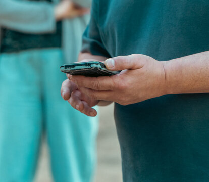A man in a dark t-shirt stands outside holding a smartphone with both hands while using modern digital technology.