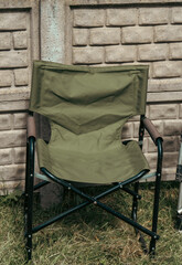 A green folding director style camping chair with a metal frame stands on grass in front of a concrete block wall. © Dmitry