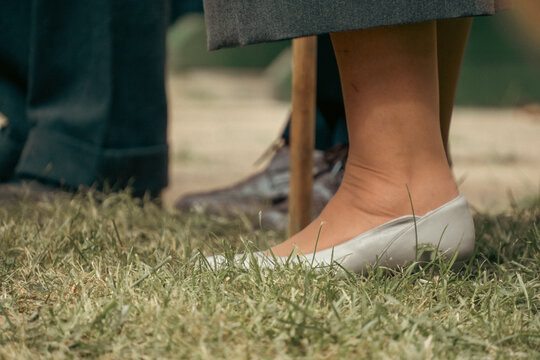 A woman stands outdoors on dry green grass wearing light grey leather shoes and a hemmed grey skirt.