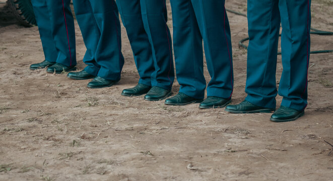 A line of officers in blue uniform trousers with red side stripes and black boots stands on the ground outdoors during a formal formation.