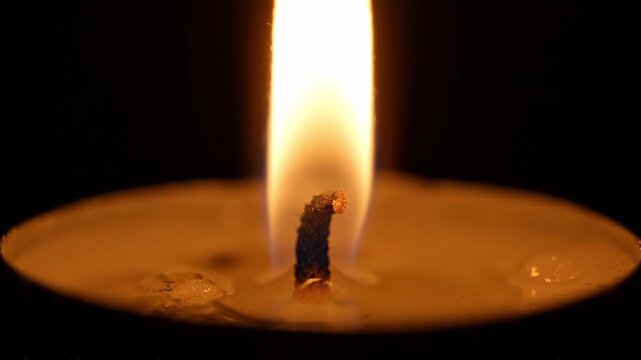 Candle flame flickers steadily as wax melts around the wick, illuminating the surrounding darkness with a warm glow in a close-up view of the candle