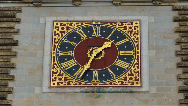 View of the ornate clock face on the Hamburg Town Hall with gold filigree, Roman numerals, and a dark red center set against a stone wall in Hamburg, Hamburg, Germany.