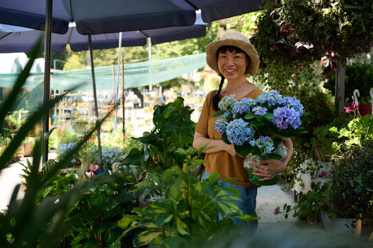 Smiling Asian Woman Holding Blue Hydrangea at Outdoor Plant Market With Straw Hat