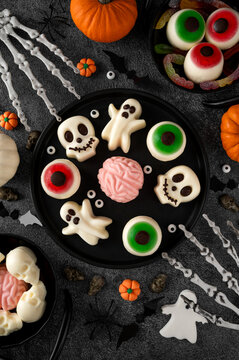Halloween jelly desserts shaped like spooky ghosts, skulls and eyeballs, served on black plates with pumpkins and festive decorations. Top view, copy space.