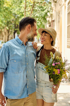 Smiling Couple Walking Arm in Arm on Sunny City Street With Bouquet and Casual Summer Clothes