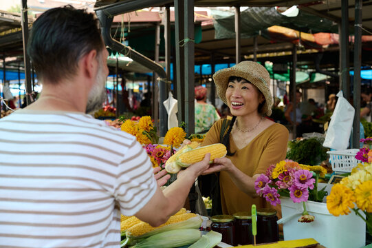 Asian Woman Vendor Smiling and Handing Fresh Corn to Customer at Outdoor Farmers Market