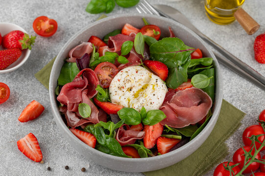 Salad with strawberries, tomatoes, burrata and jamon. Fresh summer salad in a bowl on a gray background. Top view.