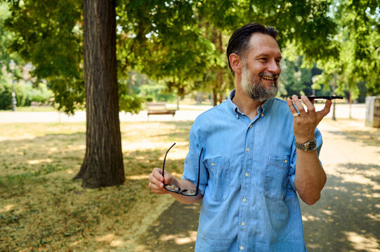 Smiling Middle-Aged Man Using Smartphone Speakerphone in Park While Holding Sunglasses