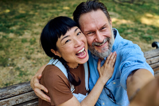Happy Middle-Aged Couple Taking Selfie On Park Bench Laughing Together Outdoors For Lifestyle