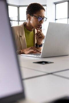 Woman typing on laptop at conference table, wearing beige blazer, eyeglasses, smartphone nearby