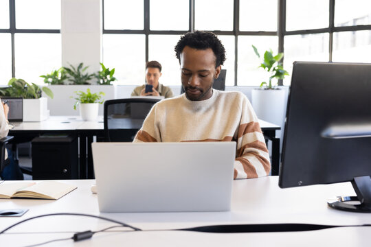 Diverse male coworkers working at open-plan office desks with laptop, monitor, notebook and plants