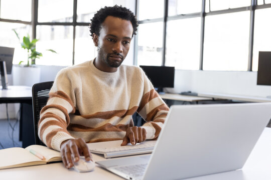 Adult African male sitting at office desk wearing cream-brown sweater using silver laptop and mouse
