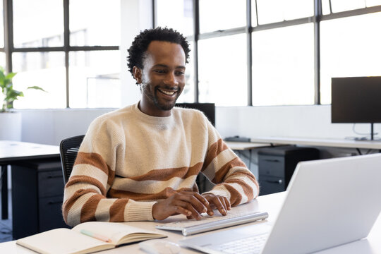 African American man smiling, typing on wireless keyboard and viewing laptop in open-plan office