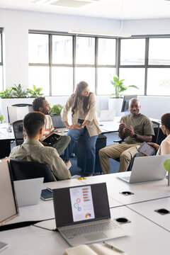 Diverse coworkers discussing project around desks in open-plan office with laptops and tablet