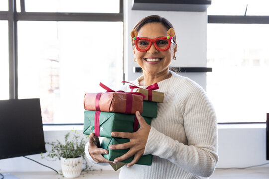 Senior African American woman wearing white knit red glasses holding three gifts at desk in office