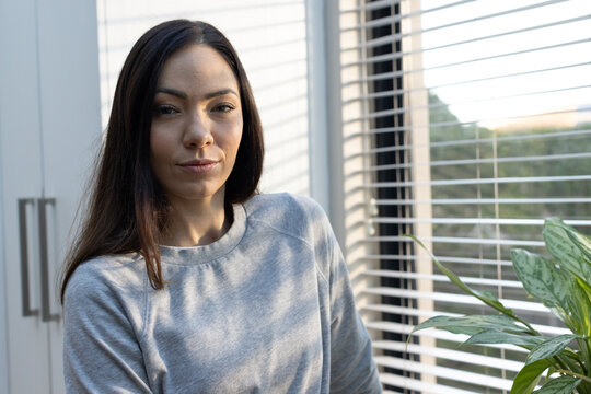 Adult woman standing by window blinds, wearing gray sweatshirt, plant and cabinet handles visible
