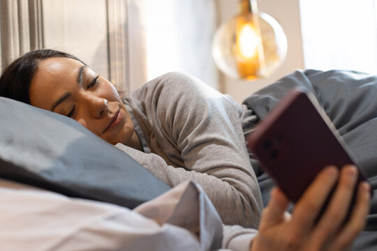 Asian woman lying on bed wearing grey top, holding purple smartphone by grey pillow under lamp
