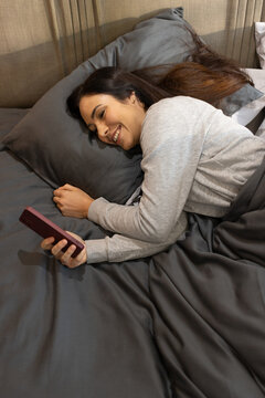 Woman lying on right side on bed holding smartphone with pink case under dark duvet