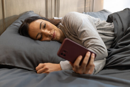 African American woman lying on side in bedroom wearing grey top holding maroon smartphone