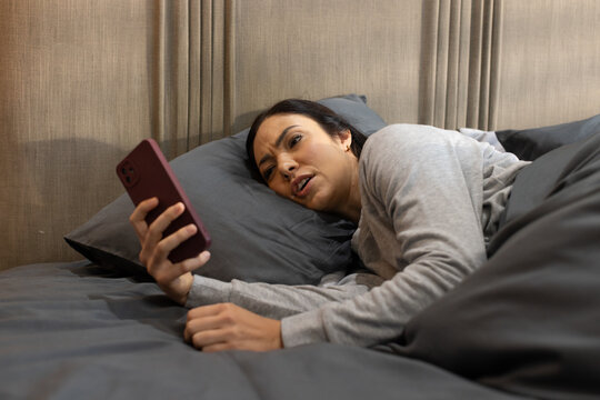 Female lying on bed on dark gray pillow holding maroon-shelled smartphone and frowning at screen