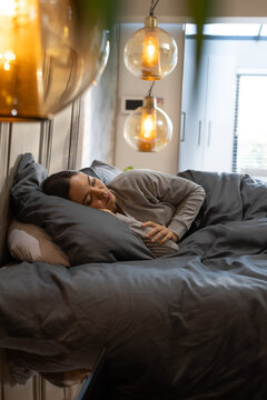 African American woman lying on made bed in bedroom wearing gray top near globe pendant lights