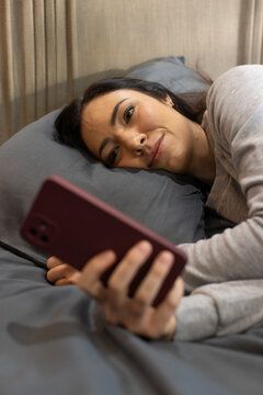 Woman reclining on bed wearing light grey top holding maroon smartphone and looking at screen