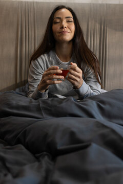 Adult woman sitting in bed holding glass of reddish tea in gray sweatshirt with dark duvet