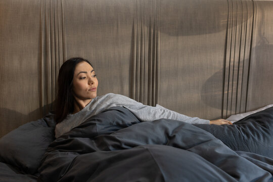 Asian woman reclining on bed in bedroom, holding pillow under dark gray duvet and headboard